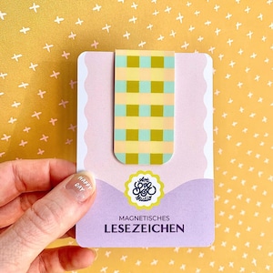 Könnte beinhalten: Ein magnetischer Lesezeichen mit einem gelb-grünen Karomuster. Das Lesezeichen befindet sich auf einer lavendelfarbenen Karte mit dem Text "MAGNETISCHES LESEZEICHEN" und einem Logo. Der Hintergrund ist gelb mit weißen Kreuzen.