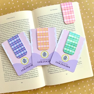 Könnte beinhalten: Vier magnetische Lesezeichen mit Vichy-Karomustern in Pink, Lila, Orange und Grün. Jedes Lesezeichen ist auf einer helllila Karte befestigt, die den Text Magnetisches Lesezeichen und ein kreisförmiges Logo Art Box Studio zeigt.