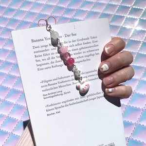Könnte beinhalten: Ein Lesezeichen mit Perlen und einem weißen Hasenanhänger am unteren Ende, gefolgt von einer Abfolge rosa und klarer Perlen in verschiedenen Schattierungen und Größen, befestigt an einem silberfarbenen Metallclip auf der Rückseite eines Taschenbuchs.