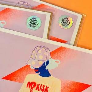 May include: A rectangular art print featuring a stylized illustration of a person wearing a polka-dot baseball cap and a cream-colored long-sleeve shirt with the red text NO RISK. The print includes a holographic sticker labeled Art Love Studio.