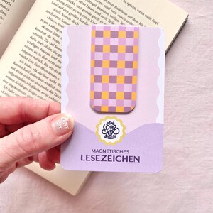 Könnte beinhalten: Ein rechteckiges magnetisches Lesezeichen mit einem violett-orangen Karomuster. Es ist auf einer hellvioletten Trägerkarte mit gewelltem Rand befestigt, die den Text Art Love Studio und MAGNETISCHES LESEZEICHEN aufweist.