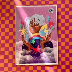 K&ouml;nnte beinhalten: Ein rechteckiger Kunstdruck mit einer stilisierten Illustration einer Person mit wei&szlig;em Haar, umgeben von bunten schwebenden Herzen, Sternen und einem Blitz. Der Hintergrund ist zartviolett mit Wolken und Glitzer, und ein silberner Aufkleber zeigt Art Soe Studio.