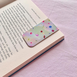 Könnte beinhalten: Ein rechteckiger Lesezeichen mit pastellgrünem Hintergrund und einer Streuung kleiner, bunter Blumen. Das Lesezeichen liegt auf einem offenen Buch mit sichtbarem Text. Das Lesezeichen verleiht dem Lesen eine florale Note.