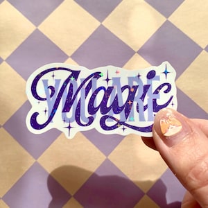 Könnte beinhalten: Ein gestanzter Aufkleber mit dem Wort Magic in einer dunkelvioletten Schreibschrift mit holografischem Sternenmuster. Hinter dem Wort Magic ist der Text YOU ARE in einer hellvioletten serifenlosen Schrift gedruckt.