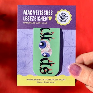 Könnte beinhalten: Ein hellgrünes magnetisches Lesezeichen mit zwei blauen Augäpfeln und dem Wort SPOOKY in schwarzen, tropfenden Buchstaben. Das Lesezeichen ist auf einer violetten und hellvioletten Trägerkarte mit Spinnennetzmuster und Text befestigt.