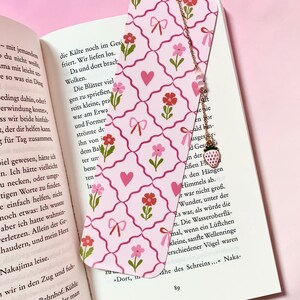 以下が含まれることがあります： ピンクの格子模様に小さな赤い花、ピンクのハート、ピンクのリボンが描かれた長方形のしおり。上部にはゴールドトーンの金属チェーンが付いており、小さなピンクのビーズとイチゴの形をしたチャームが飾られています。