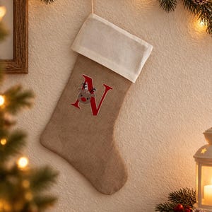 Könnte beinhalten: Ein beigefarbener Weihnachtsstrumpf aus Jute mit weißem Umschlag und Aufhängeschlaufe. Auf der Vorderseite befindet sich ein großer roter Buchstabe N, in dem eine kleine braune Rentiergrafik mit roter Nase mittig platziert ist.