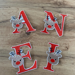 Könnte beinhalten: Vier gestickte Stoffaufnäher mit den Buchstaben A, N, E und I in einer kräftigen roten Schrift mit weißem Rand. Jeder Buchstabe ist mit einer beigen Rentierkopf-Applikation verziert, die eine leuchtend rote Nase und schwarzes Geweih hat.