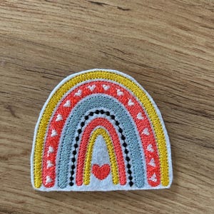 Könnte beinhalten: Ein gestickter Aufnäher in Regenbogenform auf einer weißen Filzunterlage. Das Design zeigt konzentrische Bögen in Gelb, Koralle und Hellblau, akzentuiert durch kleine weiße Herzmuster und ein einzelnes, ausgefülltes korallenrotes Herz.