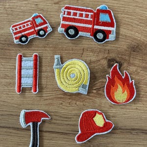 May include: A collection of seven embroidered patches featuring firefighting themes, including two red fire engines, a red ladder, a coiled yellow fire hose, a flame, a red axe with a black handle, and a red firefighter helmet with a yellow shield.