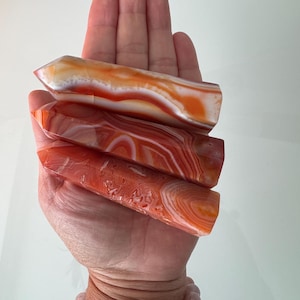 May include: Four polished agate crystal wands in shades of orange, red, and white are held in a hand. The wands have a smooth, glossy finish and display natural banding patterns. A collection of beaded bracelets is visible at the wrist.