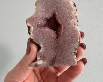 May include: A pink amethyst geode with a large opening revealing the crystal structure inside. The geode is a natural formation with a rough exterior and a smooth, polished interior.