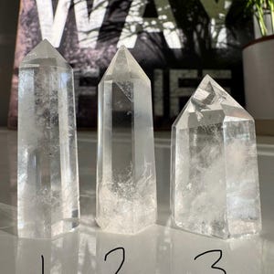May include: Three clear quartz crystal points, each with a polished, faceted design. The crystals are numbered 1, 2, and 3 with a black marker. The background features the word "WAY" in large white letters.