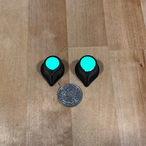 Peut inclure: Deux boutons de guitare noirs et turquoise avec un fond pointu. Les boutons sont à peu près de la taille d'une pièce de 20 centimes d'euro.