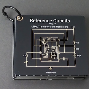 Op de afbeelding: Een stapel zwarte referentiekaarten met een metalen ring. De kaarten tonen goudkleurige schakelschema's en tekst, waaronder "Reference Circuits VOL 2" en "LEDs, Transistors and Oscillators". De naam van de auteur, Ian Dunn, is ook zichtbaar.