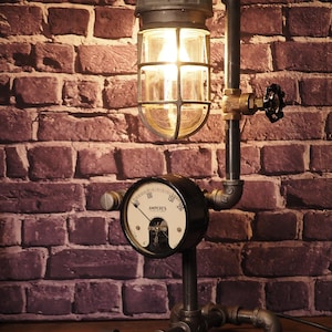 May include: Industrial-style desk lamp with a metal pipe frame and a caged glass light fixture. The lamp features a black gauge with the word "AMPERES" and a valve. The lamp is set against a brick wall.
