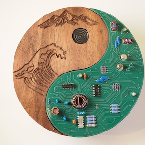 May include: A circular wall art piece shaped like a yin-yang symbol. One half is dark brown wood with laser-engraved mountain and wave designs. The other half is a green printed circuit board populated with various electronic components.