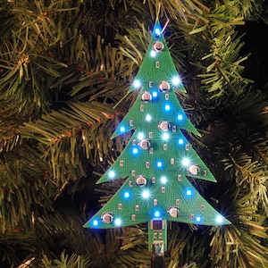 Könnte beinhalten: Eine grüne Leiterplatte in Form eines Weihnachtsbaums, die mit mehreren kleinen elektronischen Bauteilen und hellblauen Leuchtdioden auf der Oberfläche bestückt ist, mit einem kleinen Anschluss an der Basis der Baumstruktur.