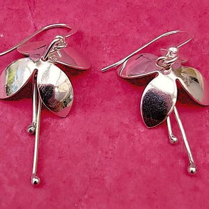 Può includere: Un paio di orecchini pendenti argentati. Ogni orecchino presenta tre elementi a forma di petalo e uno stelo pendente con una piccola sfera all'estremità. Su sfondo rosa.
