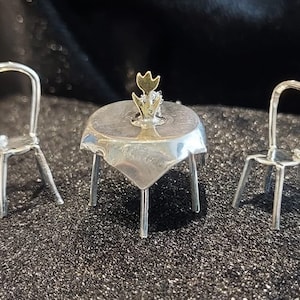 Puede incluir: Un juego de mesa de plata en miniatura con dos sillas y una sola flor de oro como centro de mesa. La mesa y las sillas están hechas de alambre y tienen un diseño delicado e intrincado.