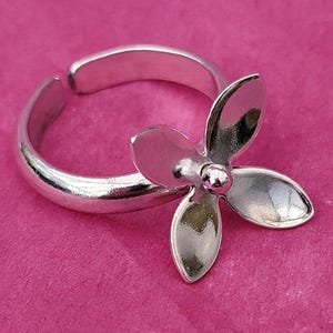 Puede incluir: Un anillo plateado con un diseño floral. El anillo presenta una flor de cuatro pétalos en el centro y una banda ajustable. La joya se muestra sobre un fondo rosa, resaltando los detalles.