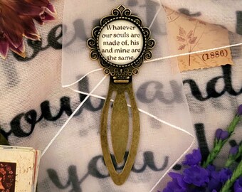 May include: A gold-toned bookmark with a round glass charm featuring the quote "Whatever our souls are made of, his and mine are the same."  The bookmark is on a white fabric background with purple flowers.