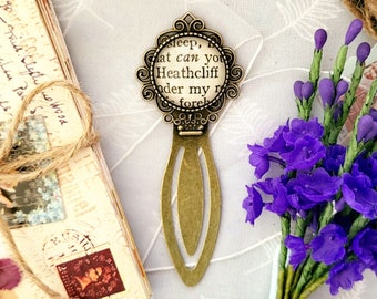 Könnte beinhalten: Ein goldenes Lesezeichen mit einem runden, gläsernen Anhänger mit dem Text "Sleep, I  that can you Heathcliff under my fore",  und einem dekorativen, verzierten Design.