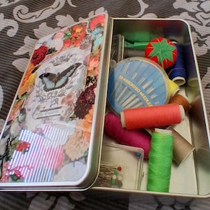 May include: Open metal sewing kit with a floral and butterfly design on the lid. The kit contains spools of thread in various colours, needles, pins, and a tomato-shaped pin cushion.