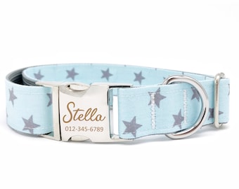 May include: A light blue dog collar with a silver buckle and a silver D-ring. The collar has a gray star pattern and the buckle has the name "Stella" and a phone number engraved on it.