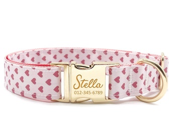 May include: A dog collar with a white background and a pattern of pink hearts. The collar has a gold-colored metal buckle engraved with the name "Stella" and a phone number. The collar is designed for pets.