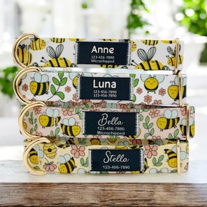 Może przedstawiać: Cztery spersonalizowane obroże dla psów ułożone w stos, każda z innym imieniem: Anne, Luna, Bella i Stella. Obroże mają wzór pszczół i kwiatów w kolorze żółtym, białym i różowym. Każda obroża ma czarną etykietę z imieniem zwierzaka i danymi kontaktowymi.