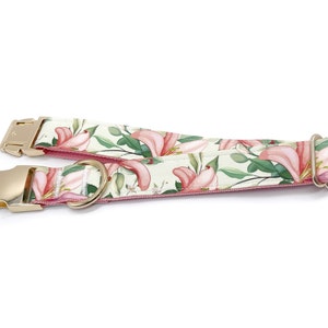 May include: A pink and green dog collar with a floral print featuring pink lilies and green leaves. The collar has a gold metal buckle and a D-ring.