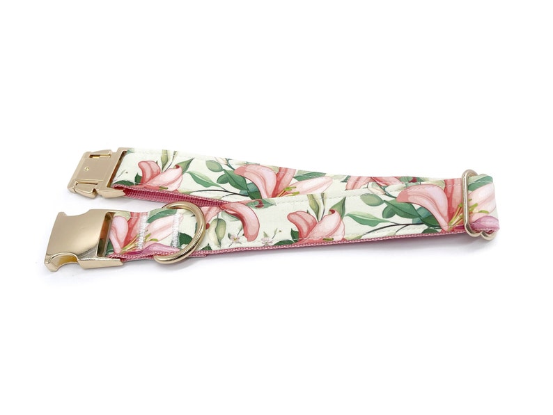 May include: A pink and green dog collar with a floral print featuring pink lilies and green leaves. The collar has a gold metal buckle and a D-ring.
