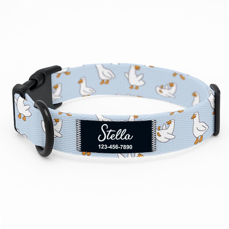 May include: Pale blue dog collar patterned with white geese. The collar has a black buckle and a black D-ring. A black rectangular patch displays the name "Stella" and a phone number.