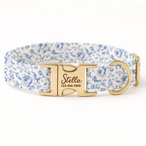 Puede incluir: Un collar para perros con un estampado floral blanco y azul, con una hebilla y una anilla en D doradas. La hebilla está grabada con el nombre "Stella" y un número de teléfono. El collar presenta un diseño floral clásico.