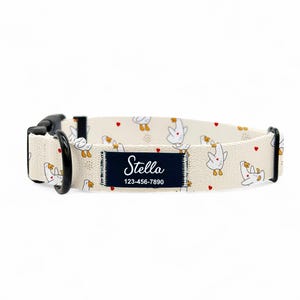 May include: A beige dog collar featuring a pattern of white cartoon geese and red hearts. The collar has a black buckle and a navy blue patch with the name "Stella" and a phone number.