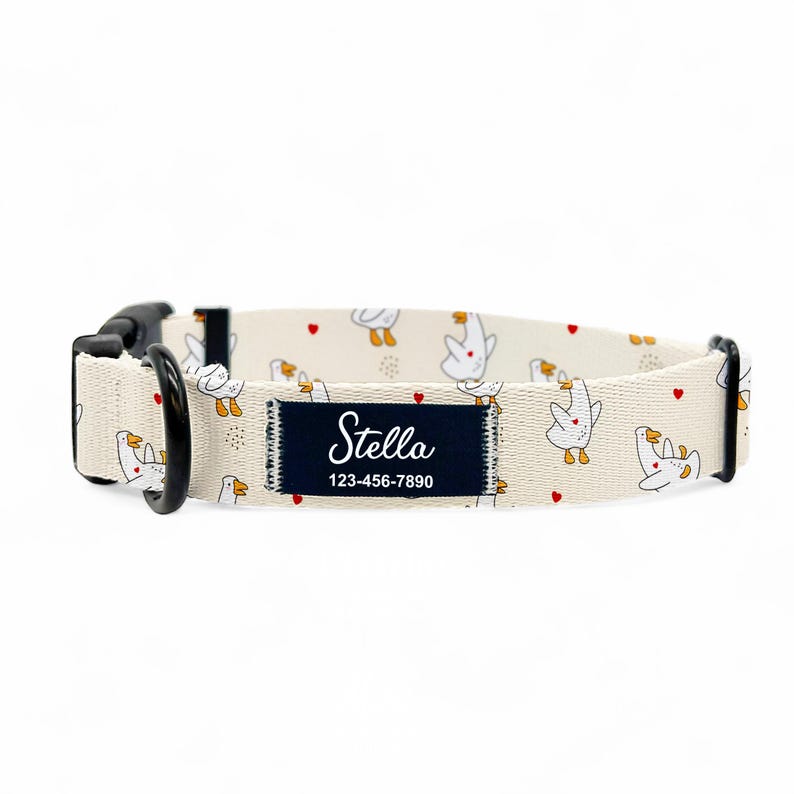 May include: A beige dog collar featuring a pattern of white cartoon geese and red hearts. The collar has a black buckle and a navy blue patch with the name "Stella" and a phone number.