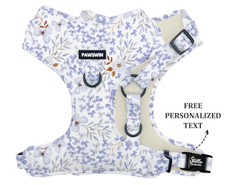 May include: White dog harness with a blue floral pattern. The harness has a mesh lining and adjustable straps. The harness has a label that says "PAWSWIN". The harness has a buckle that says "Stella".