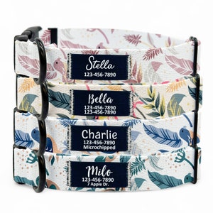 May include: Four dog collars with different names and phone numbers on them. The collars are white with a floral pattern and black hardware. The names on the collars are Stella, Bella, Charlie, and Milo.