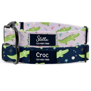 May include: Two dog collars. The top collar is light purple with green alligator graphics and the name "Stella" on a black label. The bottom collar is navy blue with green alligator graphics and the name "Croc" on a black label. Both collars have a phone number.