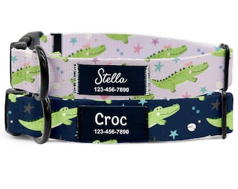May include: Two dog collars. The top collar is light purple with green alligator graphics and the name "Stella" on a black label. The bottom collar is navy blue with green alligator graphics and the name "Croc" on a black label. Both collars have a phone number.