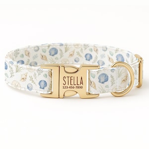Può includere: Un collare per cani con un motivo a conchiglie blu, beige e verdi su sfondo bianco. La fibbia in metallo dorato riporta il nome "STELLA" e un numero di telefono.