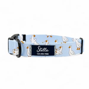 May include: A light blue dog collar featuring a pattern of white geese. The collar has a black buckle and a dark blue label with the name "Stella" and a phone number. This pet collar is designed for dogs.