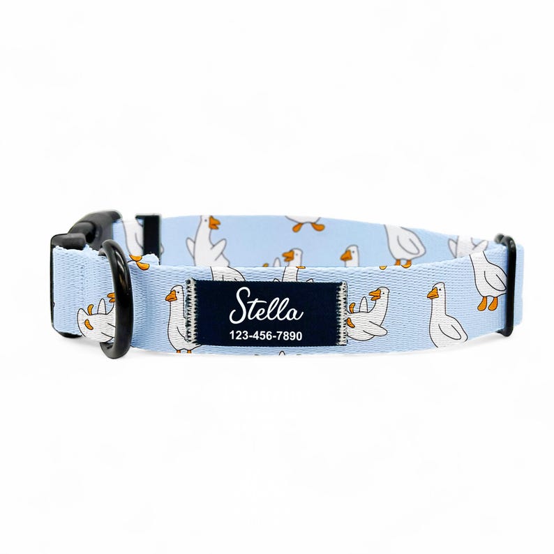 May include: A light blue dog collar featuring a pattern of white geese. The collar has a black buckle and a dark blue label with the name "Stella" and a phone number. This pet collar is designed for dogs.