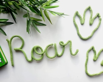 May include: Green knitted word "Roar" and two dinosaur footprint shapes on a white surface. Green leafy plant in the background. A green box with a mouse illustration is visible.