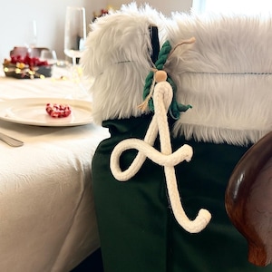 May include: A Christmas stocking with a white faux fur trim and a dark green body. A white rope initial "A" is attached with green rope and a wooden bead. The stocking is on a chair near a table setting with a white tablecloth.