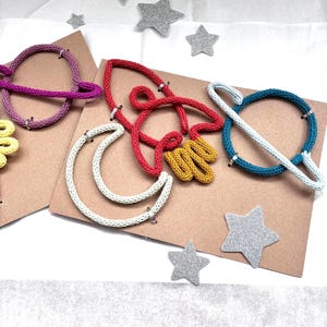 May include: A collection of colorful, handmade space-themed decorations. The set includes knitted planet and moon shapes in various colors, such as purple, red, yellow, and blue, along with silver glitter star accents. The items are arranged on brown cardstock.