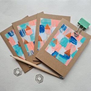 May include: Set of four blank greeting cards featuring abstract geometric designs in shades of blue, pink, and white with gold accents. The cards are on a white surface.