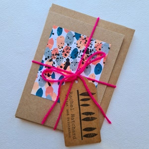 May include: A set of brown envelopes tied with a pink ribbon and a brown tag that says "Rachel Hatchard Artist and Printmaker". The envelope has a colourful abstract design with blue, pink, and grey circles.