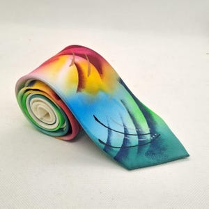 May include: A necktie rolled into a spiral, featuring a vibrant airbrushed design with gradients of magenta, yellow and turquoise. The fabric displays abstract curved black lines and soft colour transitions across the surface of the material.